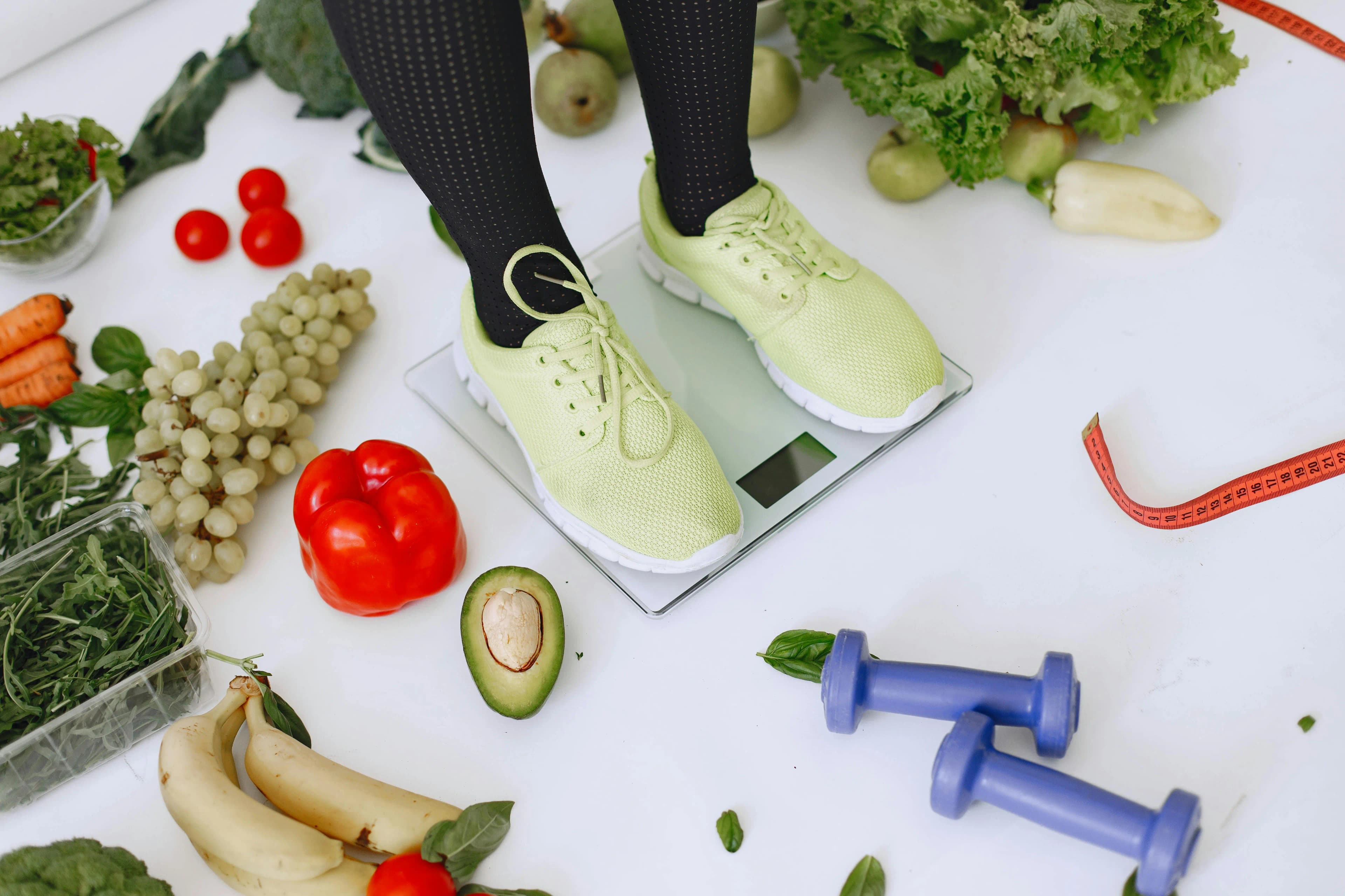 Person stepping on a scale surrounded by fresh vegetables, fruits, and dumbbells, representing Mind & Body Infusions' medically supervised medication weight loss program